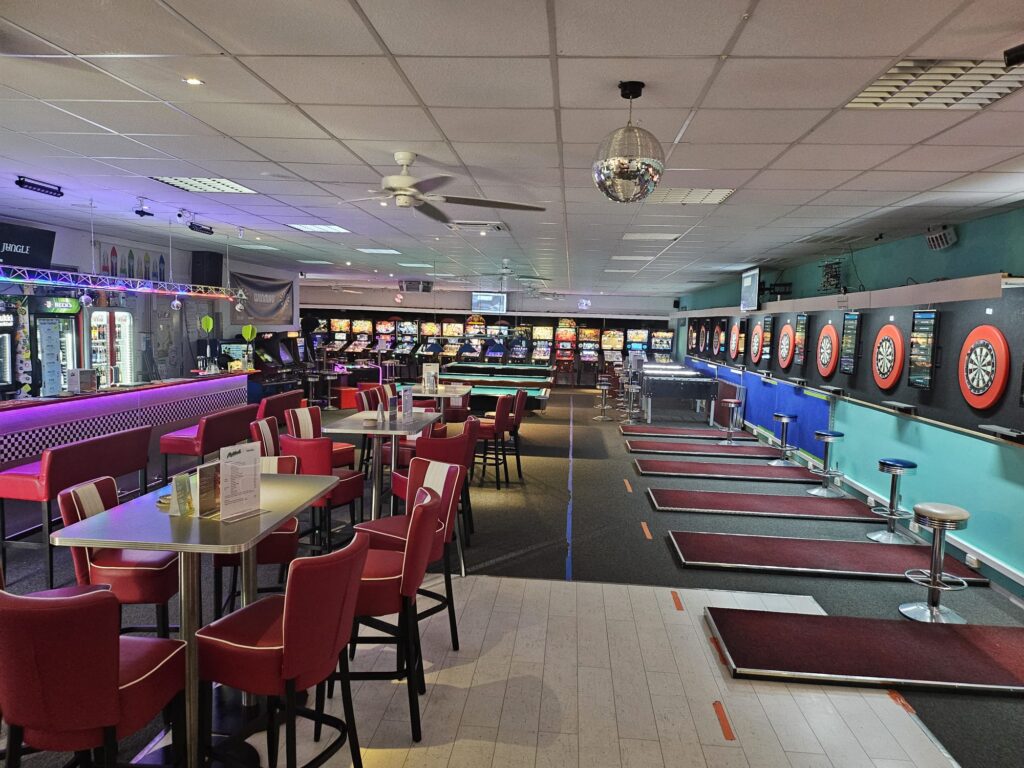 Spielhalle mit Spielautomaten, Dartboards und roten Stühlen; moderne Beleuchtung, keine Personen.