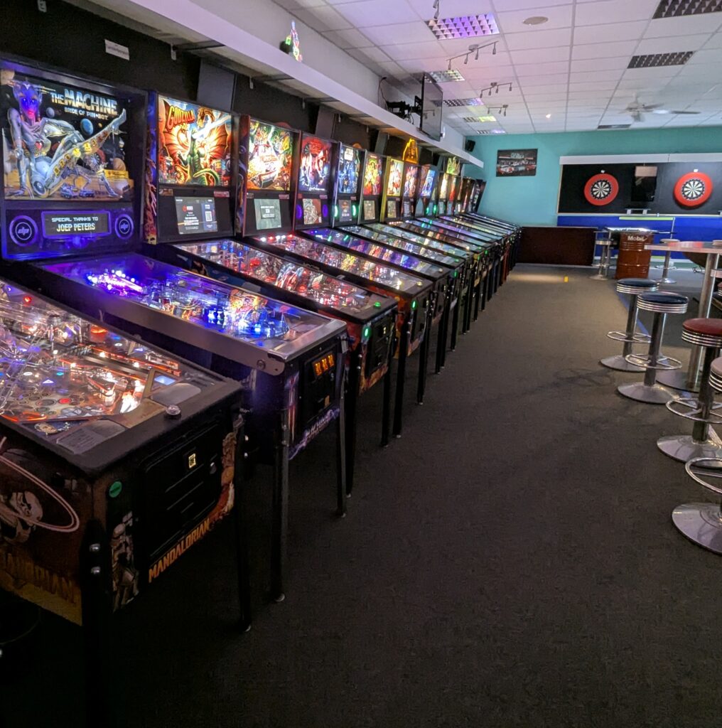 Reihe von beleuchteten Flipperautomaten in einem Arcade-Lokal mit Barhockern; farbenfrohe Lichter.