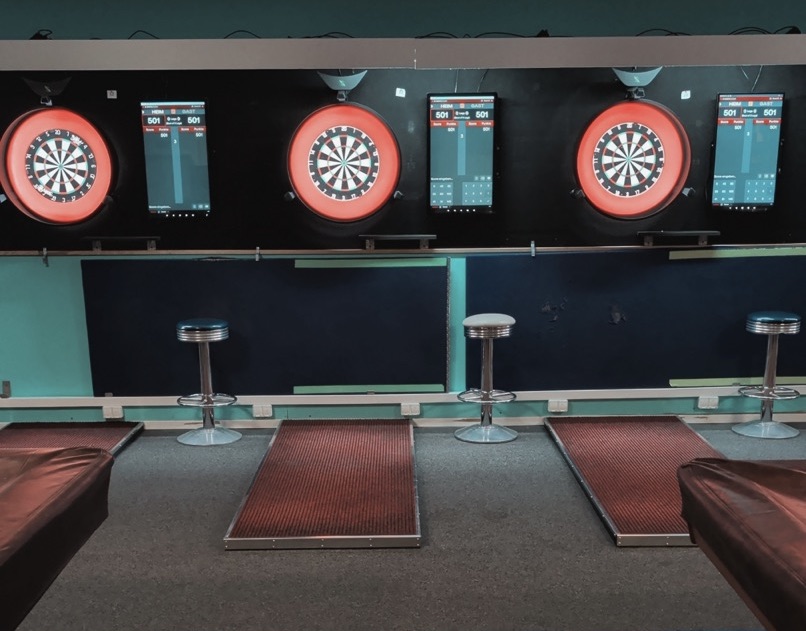 Vier elektronische Dartboards mit roten Rahmen und Bildschirmen beleuchten die Wand