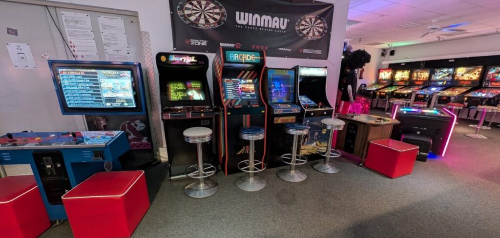 Spielhalle mit vier Arcadeautomaten in Neonfarben und roten Hockern