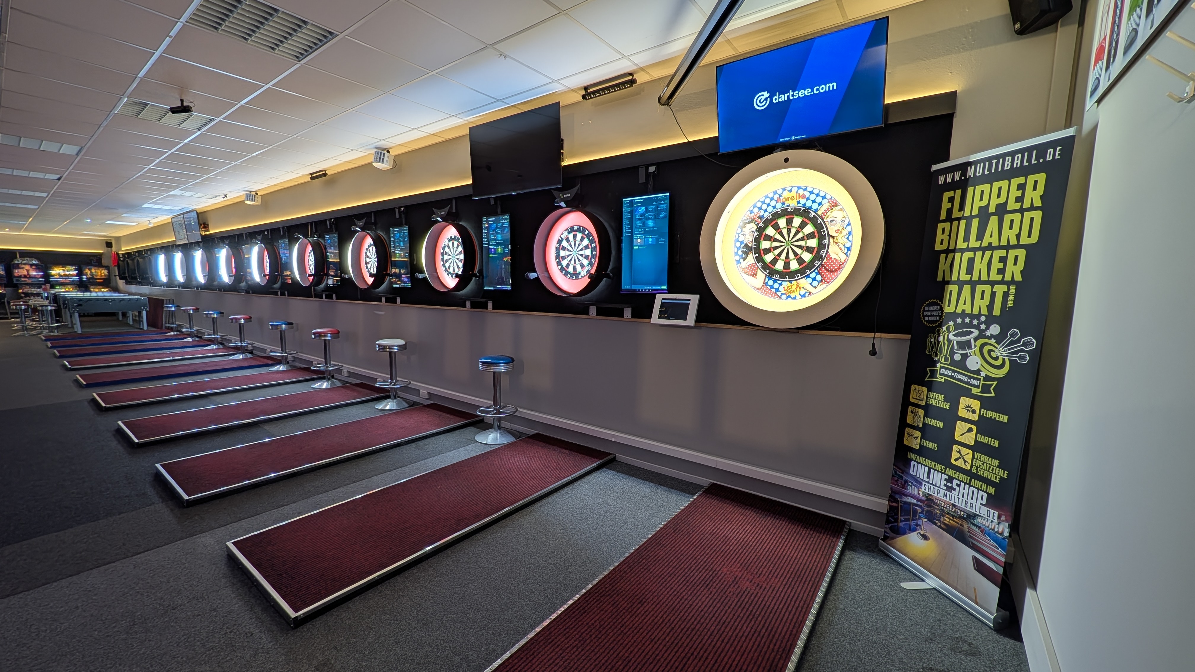 Reihe beleuchteter Dartscheiben mit Barhockern und Werbeplakat in einer Spielhalle